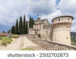 Flags flying atop the main tower of Brescia Castle in Italy, featuring a medieval drawbridge, stone walls, and the Torre dei Prigionieri on the right, with tall cypress trees along a cobblestone path