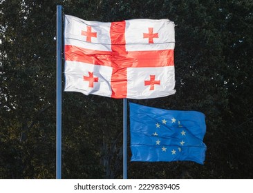 Flags of Georgia and the European Union fly in the wind in Europe Square in Tbilisi, with blurred tree foliage in the background. - Shutterstock ID 2229839405