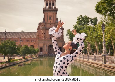 Flamenco Dancer Woman, Beautiful Brunette Teenager Dressed In Typical Costume With Ruffles And Polka Dots Is Dancing By A Canal In A Square In The Park. Flamenco Concept Of World Cultural Heritage.