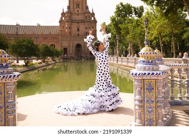 Flamenco Dancer Woman, Beautiful Brunette Teenager Dressed In Typical Costume With Ruffles And Polka Dots Is Dancing By A Canal In A Square In The Park. Flamenco Concept Of World Cultural Heritage.