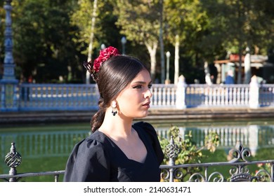 Flamenco Dancer Woman, Brunette And Beautiful Dressed In Typical Black Costume With Yellow Polka Dots Is Sitting On A Park Bench. Flamenco Concept Of Cultural Heritage Of Humanity.