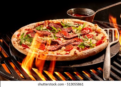 Flames Reaching Up Toward Meat And Vegetable Cheese Pizza On Grill. Hot Sauce In Bowl On Iron Plate.