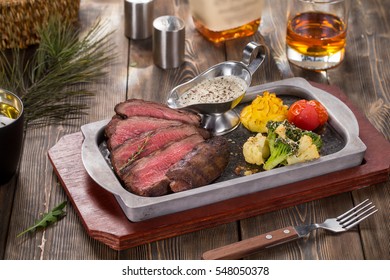 Flank Steak With Bbq Vegetables In Pan On Wooded Rustic Background With Bottle And Glass Of Whiskey In Soft Focus.