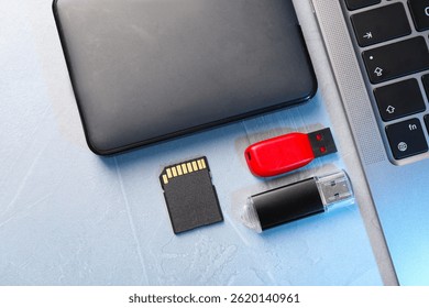 Flash drives, SD memory card, external hard disk and laptop on light grey table, flat lay