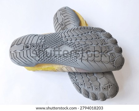 Flat angled shot of a pair of yellow barefoot shoes arranged sole-up in a crossed overlapping position on a clean white surface. Both shoes display the detailed gray rubber outsole with hexagonal tread sections, diamond grid zones, and individual toe shaped rubber pods visible on the foreground shoe. The clean white background and crossed arrangement of this barefoot shoe pair suits footwear product, sport, and minimalist shoe commercial content