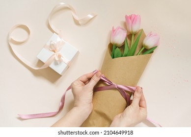 Flat Female Hands Tie A Satin Ribbon Bow On A Simple Bouquet Of Fresh Pink Tulip Flowers In Craft Paper On A Beige Table.