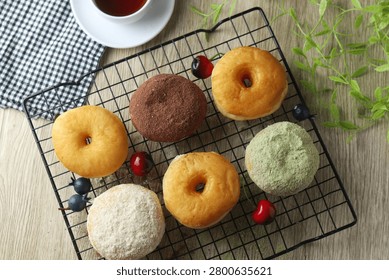Flat lay of assorted donuts on a cooling rack with cherries, berries, white tea cup, checkered napkin and green foliage on wooden table. Rustic bakery breakfast scene.
