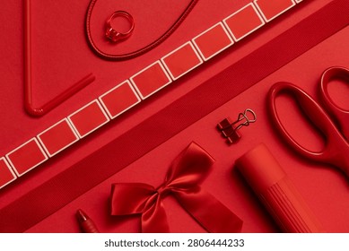 Flat lay of assorted red office and stationery supplies arranged on a matching red background. Monochromatic composition featuring scissors, ribbon, bow, pen, binder clip, flashlight, ring, and cord with ample copy space. Ideal for themes of organization, creativity, branding, education, Valentine's Day, and modern workspace design.