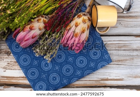 flat lay background image with Protea flowers with fynbos and enamel cups, on rustic weathered wood with unique Traditional South African printed fabric or textile. With copy space

