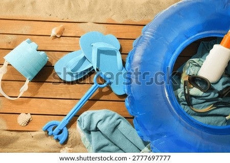 Flat lay of beach accessories including inflatable ring, flip flops, goggles, towel and toys on sand and wooden boards.