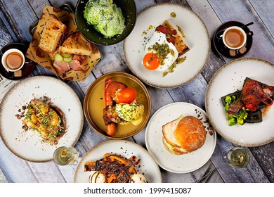 Flat lay of a beautiful brunch selection with eggs on toast, burger, pork sandwich, waffle and chocolate, salad, espresso coffee and the daily newspaper