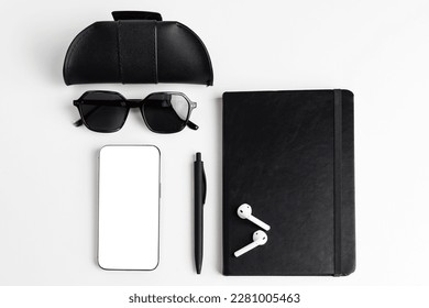 Flat lay. A black leather case for glasses, sunglasses, pen, wireless headphones, notebook, and a phone with a blank white display for copy space. Concept of organization and productivity.