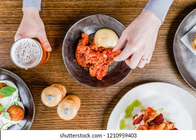 Flat Lay Of A Caucasian Hand Dipping Deep Fried Chicken In Aioli In A Black Bowl While Holding A Beer On A Wooden Table.