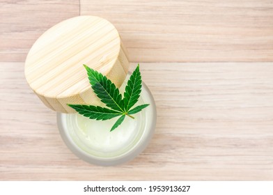Flat Lay Of CBD Lotion In Glass Container With Cannabis Leaf On Wooden Backdrop