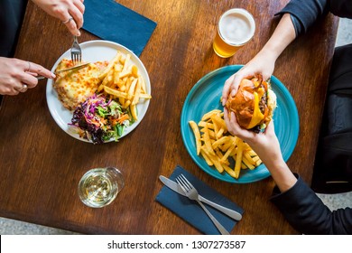 Flat Lay Of Chicken Parmigiana With Salad And Chips On A White Plate Next To An American Diner Style Burger With Fries On A Blue Plate And Glasses Of Alcohol And Caucasian Hands