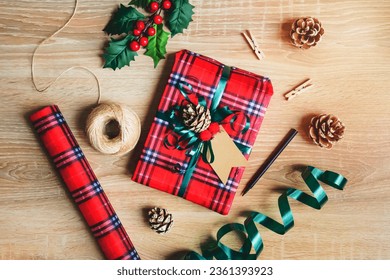 Flat lay of a Christmas gift box wrapped in scottish tartan pattern paper with wrapping materials over a wooden background. Christmas presents preparation.