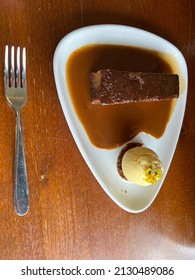 Flat Lay Close Up Of Sticky Toffee Pudding Desert, The Hot Brown Sponge Pudding Served With Thick Caramel Sauce And A Side Of Vanilla Ice Cream With Pistachio Garnish On White Plate Restaurant Lunch