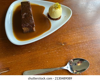 Flat Lay Close Up Of Sticky Toffee Pudding Desert, The Hot Brown Sponge Pudding Served With Thick Caramel Sauce And A Side Of Vanilla Ice Cream With Pistachio Garnish On White Plate Restaurant Lunch