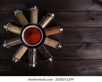 Flat lay composition of assorted ground spices in glass jars forming a circle around a white bowl filled with red pepper powder. Placed on a rustic dark wooden table with empty space for text