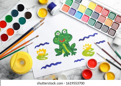 Flat lay composition with child's painting of frog on white marble table