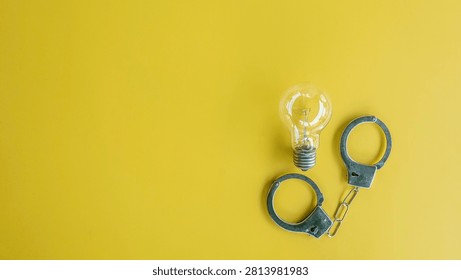 Flat lay composition of a clear glass incandescent lightbulb trapped inside a pair of open shiny steel handcuffs on a bright yellow background.