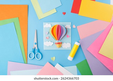 Flat lay composition with colored paper, scissors, and glue stick on a bright blue surface. DIY craft and paper art concept, kids activity, top view, craft material supply 