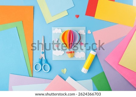 Flat lay composition with colored paper, scissors, and glue stick on a bright blue surface. DIY craft and paper art concept, kids activity, top view, craft material supply 