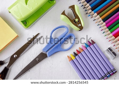 A flat lay composition featuring colorful pencils scissors a stapler and paper puncher on a clean white desk surface for back to school or office work concepts