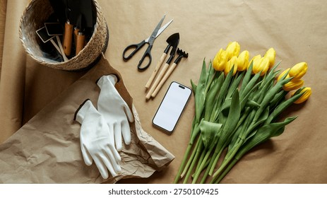 A flat lay composition featuring gardening essentials like gloves, trowel, rake, and scissors alongside a bouquet of vibrant yellow tulips and a blank screen smartphone
