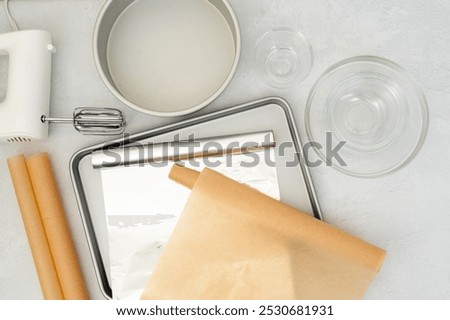 Flat lay composition featuring parchment paper, aluminum foil, a baking pan, mixer, and glass bowls arranged in a close-up view on a light grey background.