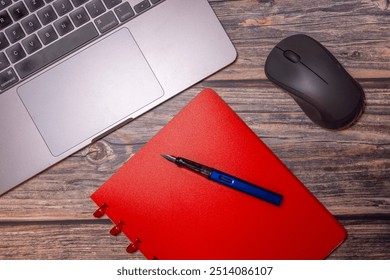 A flat lay composition featuring a red notebook, fountain pen, laptop, and mouse on a rustic wooden desk, representing modern workspace essentials for productivity and organization.