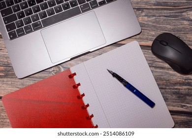 A flat lay composition featuring a red notebook, fountain pen, laptop, and mouse on a rustic wooden desk, representing modern workspace essentials for productivity and organization.