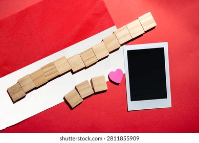 A flat lay composition featuring the red and white color, a blank instant photo frame, a small pink heart, and wooden blocks on a vibrant red background