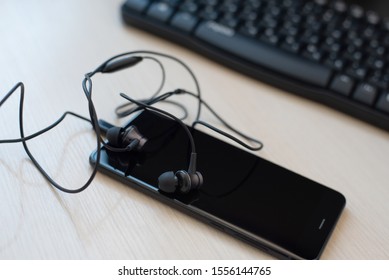 Flat lay composition with headphone and black smartphone in the foreground, against a blurred background with a computer keyboard.On begie desk background 