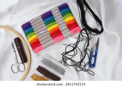 Flat lay composition of needlework tools featuring a wooden embroidery hoop, vintage scissors, a blue thread cutter, organizer box, and colorful floss skeins on white fabric.