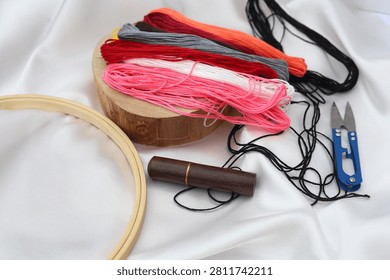 Flat lay composition of needlework tools featuring a wooden embroidery hoop, vintage scissors, a blue thread cutter, organizer box, and colorful floss skeins on white fabric.