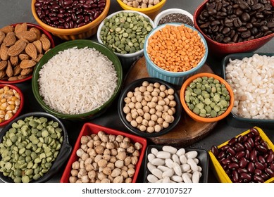 Flat lay composition showcasing a diverse assortment of healthy pantry staples. Multiple small, colorful bowls and dishes are filled with various dry ingredients including white rice, red kidney beans, almonds, green split peas, chickpeas, red lentils, soybeans, white beans, chia seeds, and corn kernels. The arrangement is set against a dark grey textured background, highlighting the different colors, textures, and shapes of these plant-based protein sources and superfoods