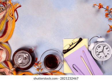 Flat lay composition showing a warm scarf, teapot, tea cup, notebooks, pen, pencil, eyeglasses, and an alarm clock on a light background, conveying an autumn learning and comfort theme