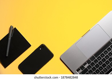 Flat lay composition with a silver grey laptop, black smartphone, notepad and a pen on an isolated yellow background.
