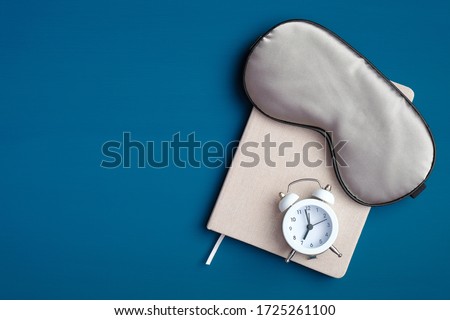Flat lay composition with sleep eye mask, dream book and alarm clock on dark blue background. Healthy sleeping concept.