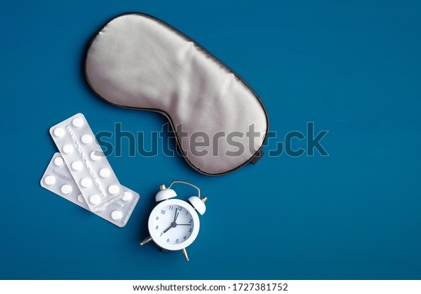 Flat lay composition with sleeping pills, sleep\
eye mask and alarm clock on dark blue background. Insomnia\
treatment, sleep healthy\
concept