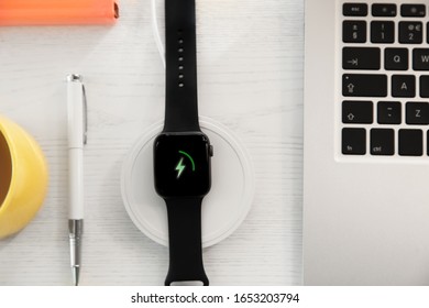 Flat lay composition with smartwatch and wireless charger on white wooden table. Modern workplace accessory