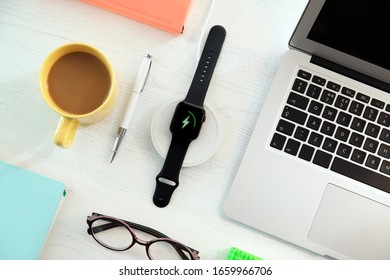 Flat lay composition with smartwatch and wireless charger on white wooden table. Modern workplace accessory