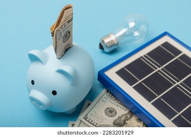 Flat lay composition with solar panel, led lamp, house model, money and piggy bank on blue background. The concept of saving money and clean energy. Concept of ecology and sustainable development.