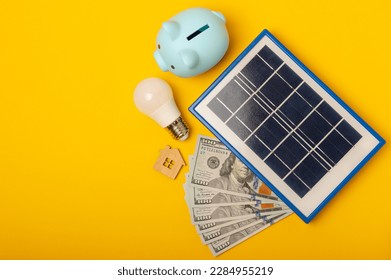 Flat lay composition with solar panel, led lamp, house model, money and piggy bank on yellow background. The concept of saving money and clean energy. Concept of ecology and sustainable development.