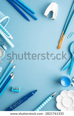 A flat lay composition of various blue school and art supplies arranged on a plain blue background, including pens, pencils, markers, scissors, paintbrushes, a sharpener, an alarm clock, and a palette. The image emphasizes organization and creativity for educational or artistic purposes.