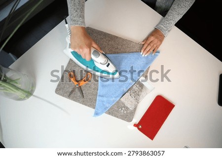 Flat lay of craft workspace featuring a person pressing blue textile next to orange scissors, pins, and a red felt pouch on a white table..JPG