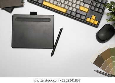 Flat lay of a creative designer's workspace featuring a drawing tablet with stylus, yellow mechanical keyboard, wireless mouse, color palette swatches, and notebook on white desk.