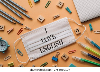 A flat lay of a creative workspace featuring a face mask with 'I LOVE ENGLISH' text, surrounded by stationery items like pens, paper clips, and a clock.