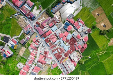 Flat lay drone shot capturing the striking visual contrast between clustered colorful rooftops of a rural settlement and vibrant green farmlands in the peri-urban area of Bukittinggi, West Sumatra, Indonesia.
